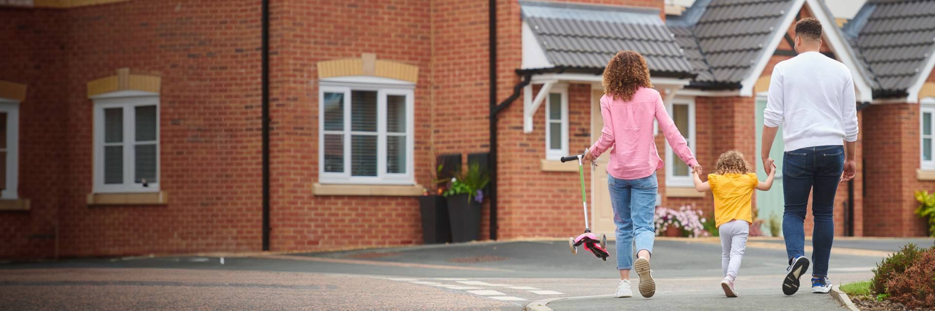 A family of three, consisting of two adults and a child, walking hand in hand down a quiet residential street. One of the adults is holding a kid scooter above ground, and they are surrounded by brick houses with white-framed windows.